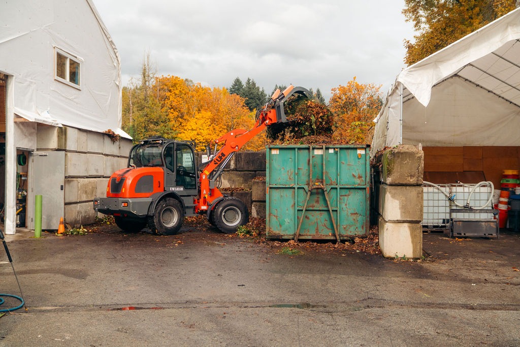 Orange loader dumping yard waste into green dumpster