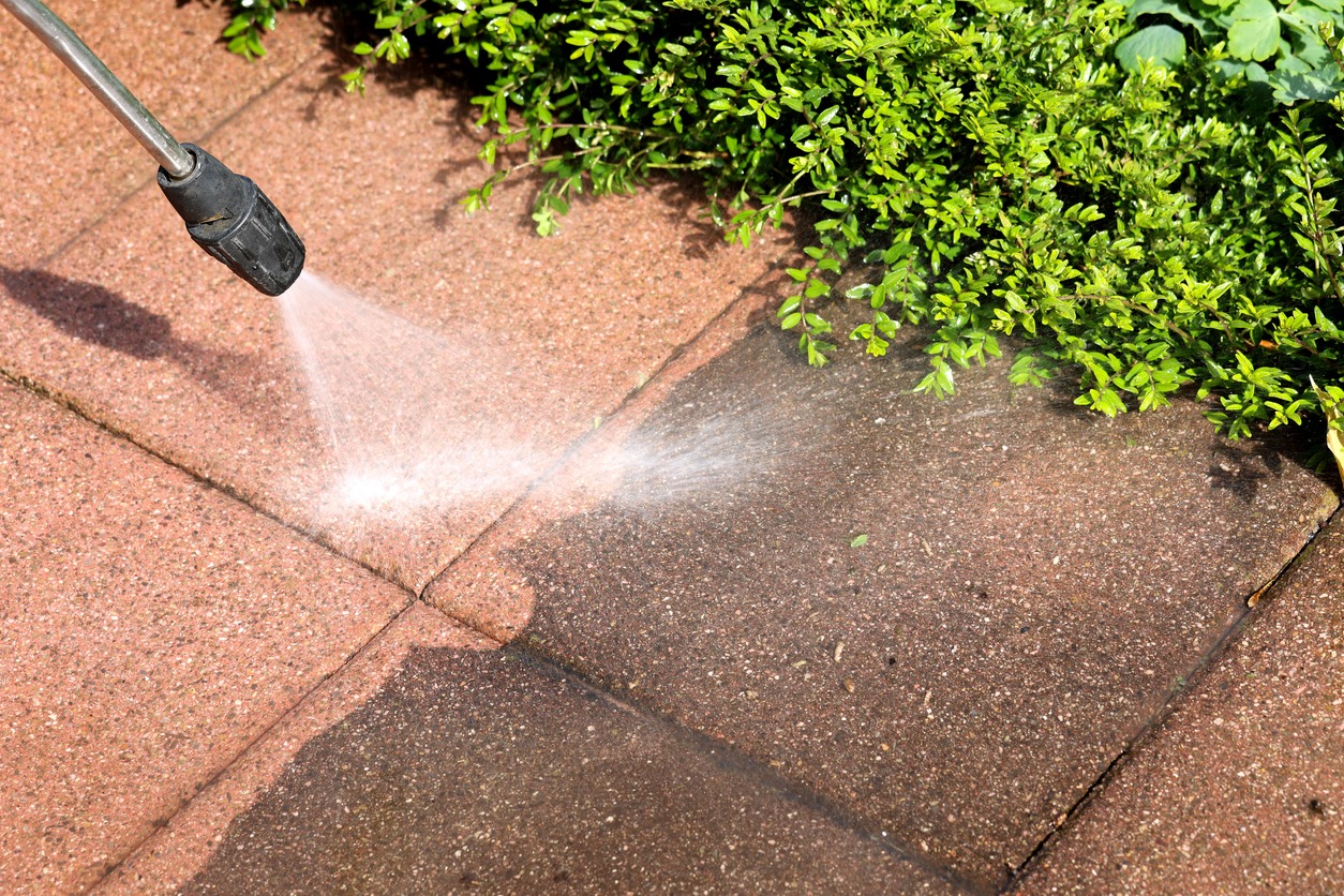 Pressure washer cleaning patio pavers near shrubs