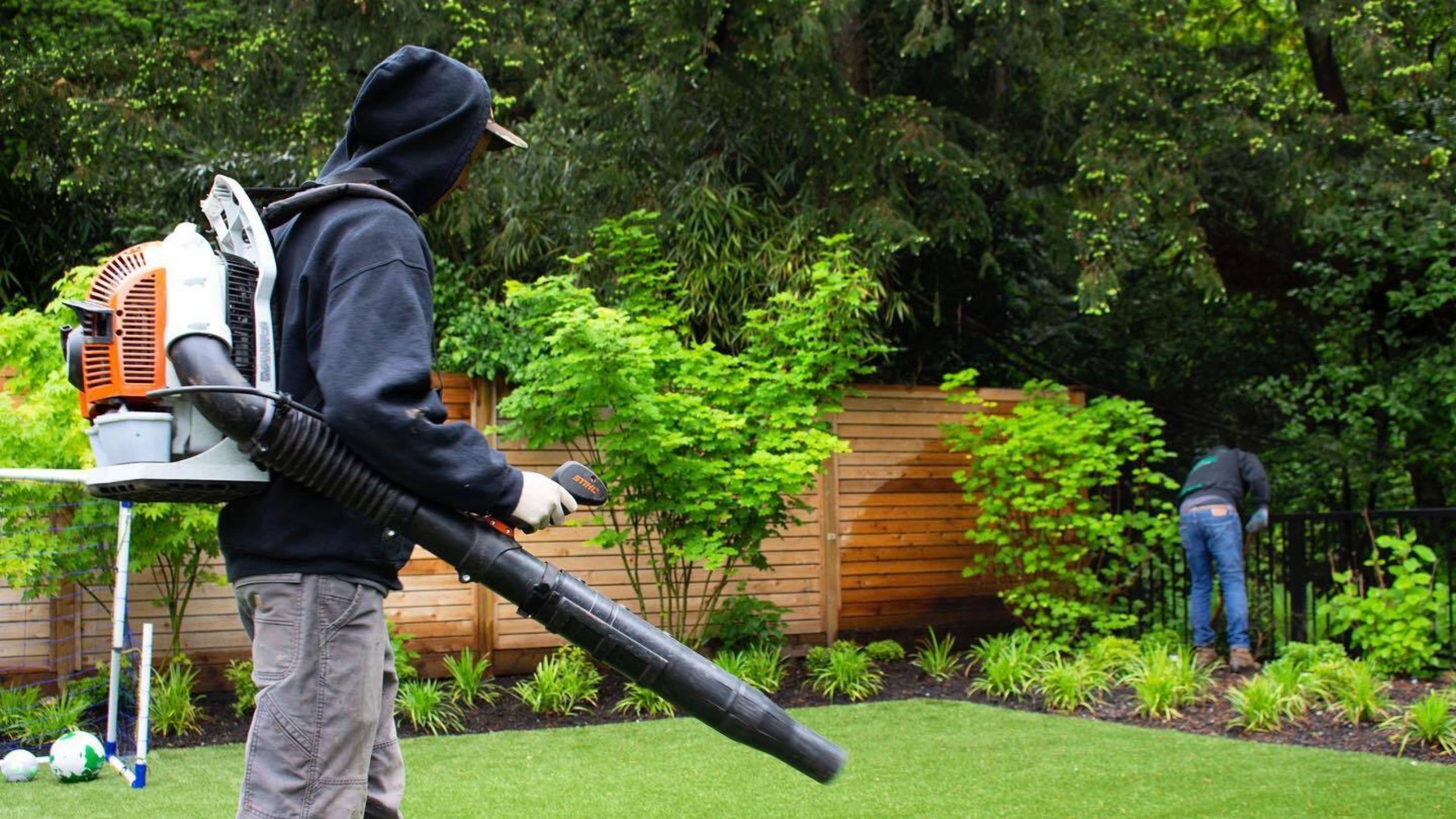 Two guys doing a yard clean up. One is holding a leaf blower and the other one is tending to shrubs.